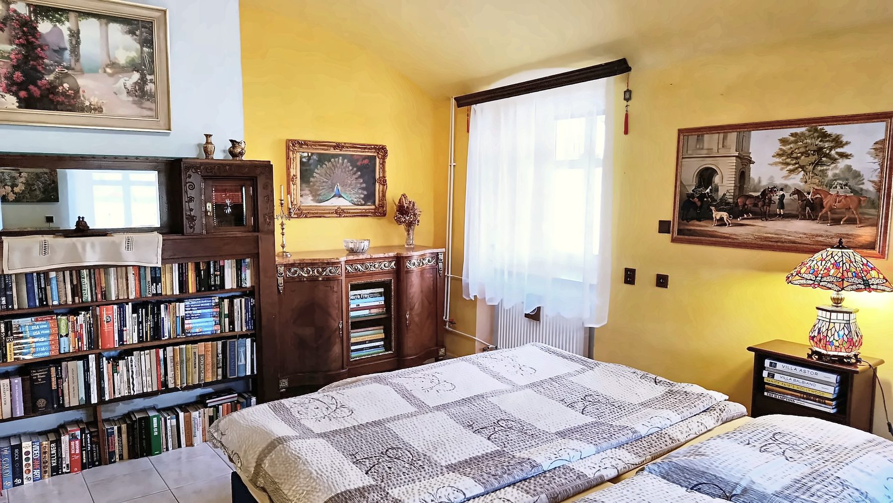 Ložnice se žlutými stěnami, manželskou postelí, knihovnou, tiffany lampou a obrazy